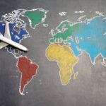 Self-Guided Tours - Top view of crop anonymous person holding toy airplane on colorful world map drawn on chalkboard