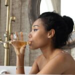 Wine Tastings - Side view of young ethnic female sitting in bathtub and drinking wine while enjoying spa procedure in bathroom