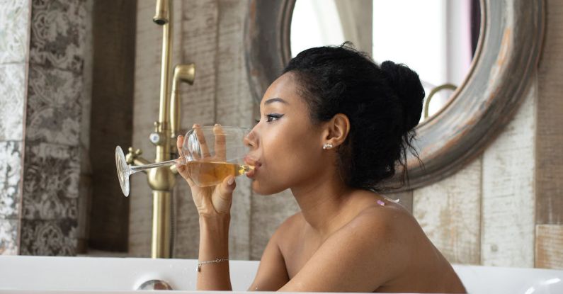 Wine Tastings - Side view of young ethnic female sitting in bathtub and drinking wine while enjoying spa procedure in bathroom